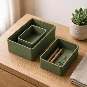 Può includere: Un set di tre vassoi portaoggetti rettangolari color verde oliva di diverse dimensioni. Un vassoio è inserito all'interno di uno più grande, mentre il terzo vassoio contiene due penne in legno e diverse graffette in metallo argentato.