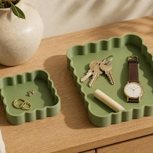 Può includere: Due vassoi quadrati color verde salvia con bordi smerlati. Il vassoio più piccolo contiene due anelli dorati. Il vassoio più grande contiene un mazzo di chiavi in metallo, un tubetto bianco cilindrico e un orologio con cinturino in pelle marrone.