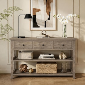 May include: A rectangular wooden console table with three drawers and two lower shelves. The table features a weathered gray finish and black hardware. It is styled with a black desk lamp, a glass vase of white orchids, books, and woven storage baskets.