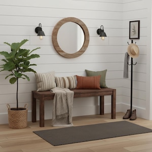 May include: A cozy entryway scene featuring a wooden bench with pillows and a throw blanket. A round wooden mirror hangs above the bench, flanked by black sconces. A coat rack holds a hat and scarf, and a potted plant adds greenery. A dark rug completes the space.