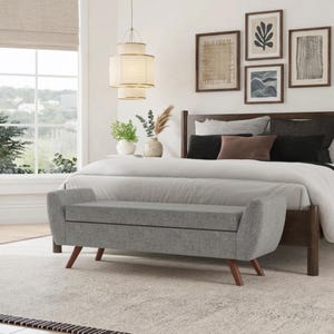 May include: A light gray upholstered bench with curved arms and tapered wooden legs positioned at the foot of a bed. The bench features a rectangular seat cushion and a solid base, providing a functional seating area in a bedroom setting.