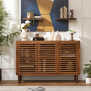 May include: A rectangular wooden sideboard featuring three cabinet doors with horizontal slats. The piece stands on four tapered legs and has a warm brown finish. The top surface holds a white pitcher with flowers, two white vases, and a fruit bowl.