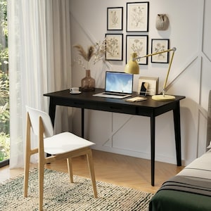 May include: A black rectangular desk with two drawers, a white chair with light wood legs, a laptop, a yellow desk lamp, a glass vase with dried plants, a small white cup, and a document holder, positioned against a wall with five framed botanical prints.