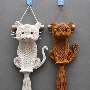 Puede incluir: Dos tapices de macramé con forma de gato, uno en color blanco roto y otro en marrón dorado. Cada gato presenta un cuerpo tejido con textura, ojos oscuros, bigotes rígidos, una cola curva y una borla larga en la base, colgados de un palo de madera.