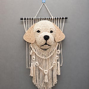 May include: A macrame wall hanging featuring a three-dimensional cream-colored dog head with tan ears, black eyes, and a black nose. The dog is mounted on a black horizontal rod with cream-colored fringe, decorative knotted loops, and tassels.