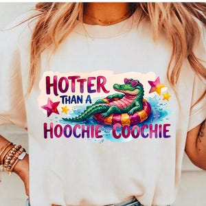 May include: A cream-colored t-shirt featuring a graphic of a green alligator wearing heart-shaped sunglasses while lounging on a pink and yellow striped pool float. The text reads "HOTTER THAN A HOOCHIE COOCHIE" in purple and pink lettering.