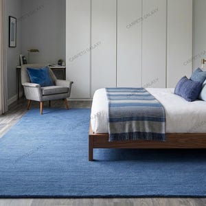 May include: A large, solid blue area rug with a textured surface covering the floor of a bedroom. The rug is positioned beneath a wooden bed frame and extends across the room, featuring a deep blue color that complements the surrounding decor.