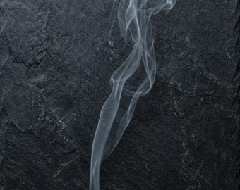 May include: A thin, wispy trail of white smoke rises vertically against a dark, charcoal-colored slate background. The surface features a rough, uneven, and layered texture with natural fissures and jagged edges characteristic of natural stone.