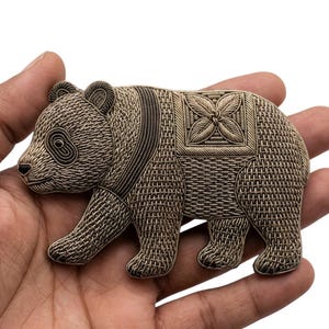 Pode incluir: Um emblema de panda em tom dourado metálico com bordado intrincado semelhante a arame, um padrão texturizado de hachuras cruzadas no corpo e um motivo floral quadrado decorativo nas costas, com costuras detalhadas ao redor dos olhos e orelhas.