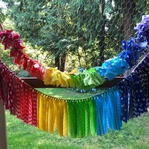 May include: A rainbow fabric garland with red, yellow, green, blue, and purple fabric strips. The garland is strung on a white string and is hanging from a wooden post.