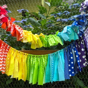 May include: A rainbow fabric garland with red, orange, yellow, green, blue, indigo, and purple fabric strips. The fabric strips are tied together with white string and have different patterns, including polka dots, stripes, and chevron.