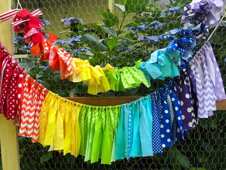 May include: A rainbow fabric garland with red, orange, yellow, green, blue, indigo, and purple fabric strips. The fabric strips are tied together with white string and have different patterns, including polka dots, stripes, and chevron.