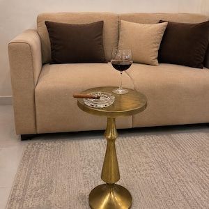 Op de afbeelding: Een ronde bijzettafel van metaal met een messing afwerking en een voetstuk. Op het tafelblad staat een asbak van helder glas met een bruine sigaar en een wijnglas gevuld met rode wijn. De tafel heeft een glad, cirkelvormig tafelblad.
