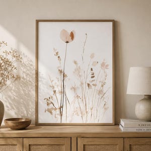 Könnte beinhalten: Ein gerahmter vertikaler Wandkunstdruck mit einer Aquarellillustration zarter Wildblumen in Beige-, Hellbraun- und Sandtönen. Das Kunstwerk ist in einem dünnen Holzrahmen gefasst und zeigt eine einzelne markante Blume sowie verschiedene Gräser.