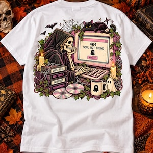 May include: A white t-shirt featuring a graphic of a skeleton in a hooded cloak sitting at a computer. The screen displays 404 SOUL NOT FOUND. The design includes a black cat, books labelled GHOST FILES, LOST SOULS, INTERNET, PARANORMAL, CDs, and a ghost mug.