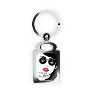 May include: A silver-toned metal keychain featuring a rectangular pendant with a graphic illustration of a face wearing round sunglasses with green leaf motifs in the lenses, red lips, and the text "Let's get SmoKEY" printed in black script.