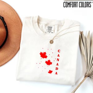 May include: A cream-colored short-sleeve t-shirt with a Comfort Colors brand label at the neck. The front features three red maple leaf graphics of varying sizes, small red star accents, and the vertical red text reading CANADA.