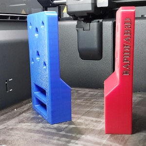 Pode incluir: Dois objetos impressos em 3D: um suporte azul com furos e uma peça retangular vermelha com o texto "RAPIDEAB3D". Ambos estão em uma base de impressora. O suporte azul tem duas prateleiras e vários furos circulares. A peça vermelha é sólida.