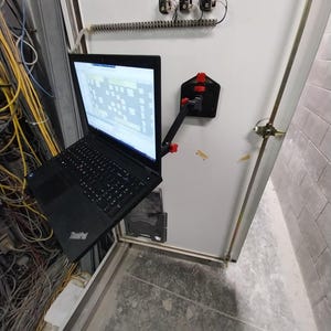 May include: A black laptop mounted on a white cabinet door using an adjustable black metal arm with red knobs. The laptop screen displays a grid-like interface. The arm is secured to the door with a black triangular base plate and red fasteners.