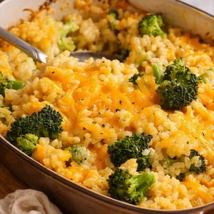 May include: A baked casserole dish filled with a mixture of cooked rice, bright green broccoli florets, and melted orange cheddar cheese. A metal serving spoon is partially submerged in the food, which is seasoned with visible black pepper flakes.