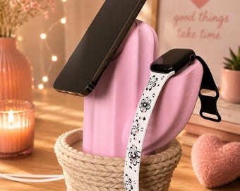 May include: A pink cactus-shaped charging station with a beige woven-style base. The device holds a black smartphone at an angle and a smartwatch with a white floral-patterned band. The structure is designed to support and charge electronic devices.