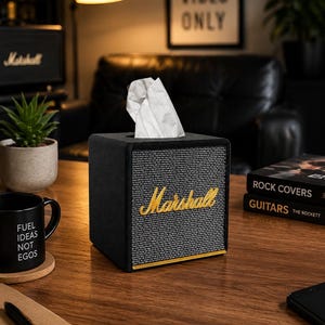 Puede incluir: Una caja de pañuelos cuadrada de color negro diseñada para parecer un amplificador de guitarra vintage. El frente presenta una rejilla de tela gris texturizada con el logotipo Marshall en letras doradas y un pañuelo blanco sobresaliendo.