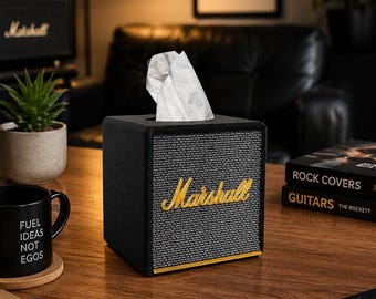 May include: A black square tissue box cover designed to resemble a vintage guitar amplifier. The front features a grey textured grille cloth with the gold script logo Marshall centered, and a white tissue protruding from the top opening.