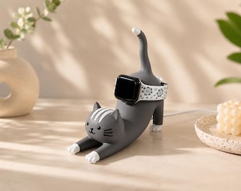 May include: A dark grey cat-shaped charging stand for a smartwatch. The cat is in a stretching pose with white paws and white stripes on its head. A black smartwatch with a white floral patterned band is wrapped around the cat's midsection.