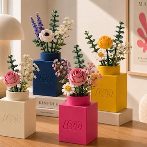 Puede incluir: Cuatro jarrones cuadrados LEGO en crema, azul, rosa y amarillo, cada uno con el logotipo de LEGO en relieve. Estos recipientes contienen varios arreglos de flores artificiales, incluyendo rosas rosas, rosas amarillas, margaritas y lavanda.