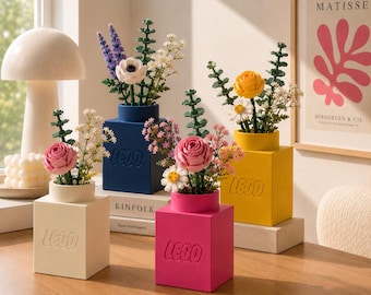 May include: Four square LEGO vases in cream, dark blue, hot pink, and yellow, each embossed with the LEGO logo. These vases hold various plastic flower arrangements, including pink roses, white anemones, yellow daisies, and green foliage stems.