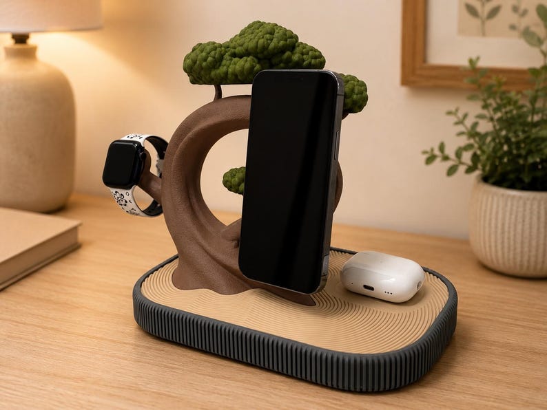 May include: A multi-device charging station featuring a brown tree-shaped stand with green foliage. The base has a tan, textured surface with circular patterns and a dark grey ridged edge. It holds a black smartphone, a smartwatch, and white earbuds.