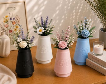 May include: Four conical vases in black, white, pink, and light blue, each embossed with the LEGO logo. Each vase contains a unique arrangement of plastic botanical flowers, including roses, daisies, lavender, and sprigs of greenery.
