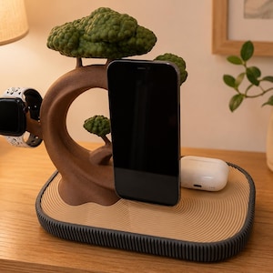 May include: A brown tree-shaped charging station with green foliage, featuring a black smartphone, a white smartwatch with a patterned band, and white wireless earbuds. The base has a tan, rippled surface and a dark grey, ribbed edge.