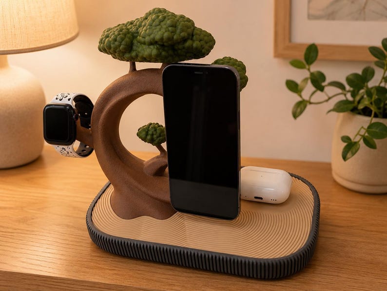 May include: A brown tree-shaped charging station with green foliage, featuring a black smartphone, a white smartwatch with a patterned band, and white wireless earbuds. The base has a tan, rippled surface and a dark grey, ribbed edge.
