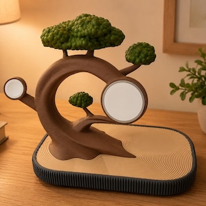 May include: A brown bonsai tree-shaped charging station with green foliage, featuring a circular white magnetic charger for a phone and a smaller circular charger for a watch, set on a tan base with a textured sand-like pattern and a dark grey rim.