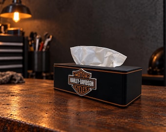 May include: A rectangular black tissue box holder featuring the orange, white, and black Harley-Davidson Motor Cycles logo on the front. The box has thin orange pinstripe detailing along the top and bottom edges and contains white facial tissues.