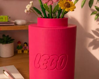 May include: A bright pink cylindrical LEGO vase featuring the embossed LEGO logo on the front. The vase is filled with a colorful bouquet of artificial flowers constructed from plastic building bricks, including white, pink, and yellow blossoms.