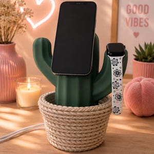 May include: A dark green cactus-shaped charging station base set inside a beige rope-textured basket. The cactus features a central slot holding a black smartphone and a side arm holding a smartwatch with a white band featuring black flower patterns.