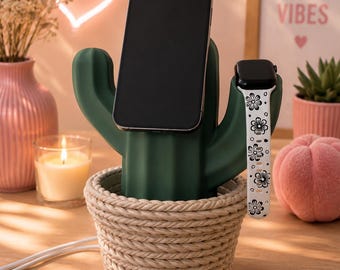 May include: A dark green cactus-shaped charging station base set inside a beige rope-textured basket. The cactus features a central slot holding a black smartphone and a side arm holding a smartwatch with a white band featuring black flower patterns.