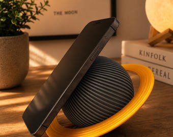 May include: A dark gray spherical phone stand with a ribbed texture and a bright yellow ring encircling its center, designed to hold a black smartphone at an angle. The stand resembles a planet with rings, providing a stable base for the device.