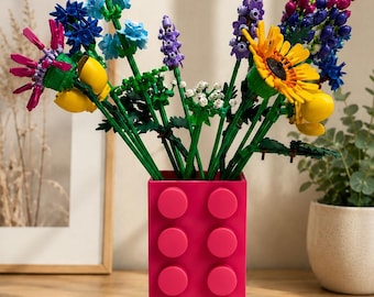 May include: A bouquet of colorful plastic building block flowers in shades of yellow, blue, purple, and pink, arranged in a rectangular, bright pink plastic vase with a textured, studded surface resembling a large building block brick.