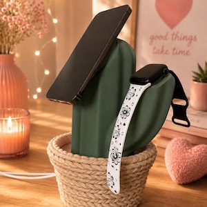 May include: A dark green cactus-shaped charging stand for a smartphone and a smartwatch. The cactus is set in a beige woven basket base. A black smartphone rests against the main cactus body, while a white watch band with black floral patterns hangs over the side arm.