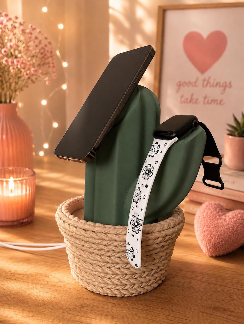 May include: A dark green cactus-shaped charging stand for a smartphone and a smartwatch. The cactus is set in a beige woven basket base. A black smartphone rests against the main cactus body, while a white watch band with black floral patterns hangs over the side arm.