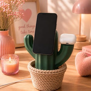 May include: A dark green cactus-shaped charging station with a woven rope base. The device features a central upright slot for a smartphone and a smaller side platform for wireless earbuds, connected by a white power cord extending from the bottom.