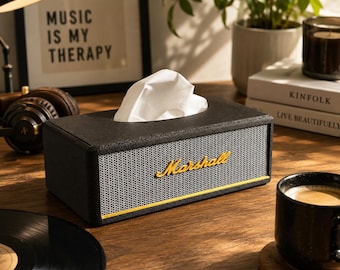May include: A rectangular black tissue box cover designed to resemble a vintage guitar amplifier. It features a textured black exterior, a grey woven front grille, a gold-colored Marshall logo, and a thin gold trim line along the bottom edge.