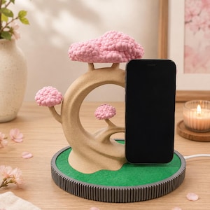 May include: A phone stand shaped like a tan tree with textured pink blossoms. The stand sits on a circular base with a green top and a dark grey ribbed edge. A black smartphone is positioned upright against the tree structure, connected by a white charging cable.