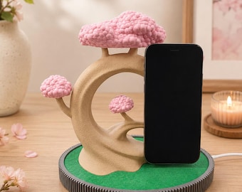 May include: A phone stand shaped like a tan tree with textured pink blossoms. The stand sits on a circular base with a green top and a dark gray ribbed edge. A black smartphone is positioned upright against the tree structure, connected by a white charging cable.