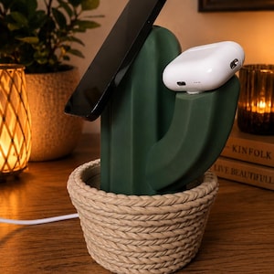 May include: A dark green cactus-shaped charging station with a beige rope-textured base. The device holds a black smartphone at an angle and a white wireless earbud case on a side arm, with a white charging cable extending from the base.