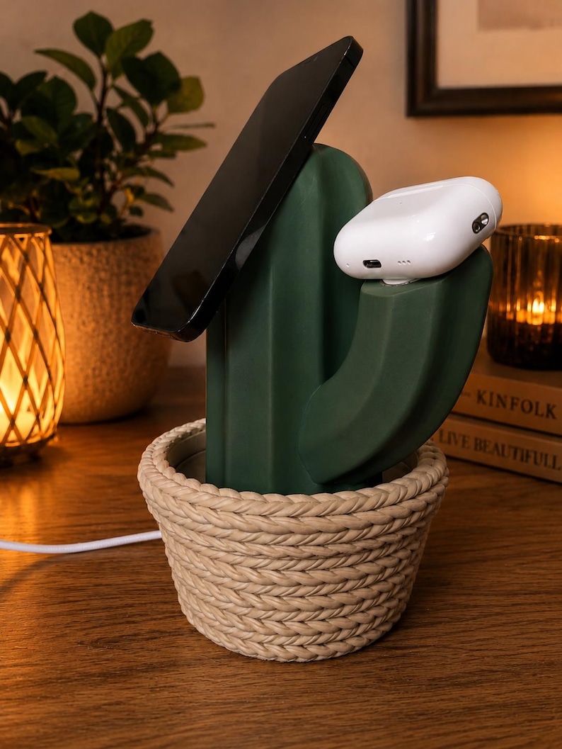 May include: A dark green cactus-shaped charging station with a beige rope-textured base. The device holds a black smartphone at an angle and a white wireless earbud case on a side arm, with a white charging cable extending from the base.