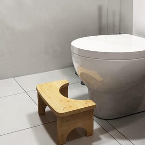 May include: A light brown bamboo toilet stool with a curved cutout design. The stool features a rectangular top with rounded corners and four sturdy legs with arched openings at the base, positioned on a white tiled floor next to a white toilet.